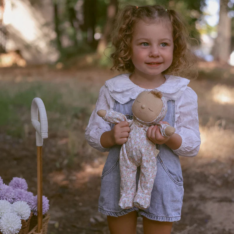 A young child happily holding a Dozy Dinkum Custard plush toy by Olli Ella in an outdoor setting. The child is smiling and wearing a stylish outfit, showcasing the toy's playful nature.