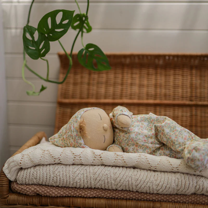 A Dozy Dinkum Custard plush toy by Olli Ella, softly lying on a knitted blanket in a cozy setting. The plush has a floral print and is surrounded by natural decor, enhancing its comfy vibes.