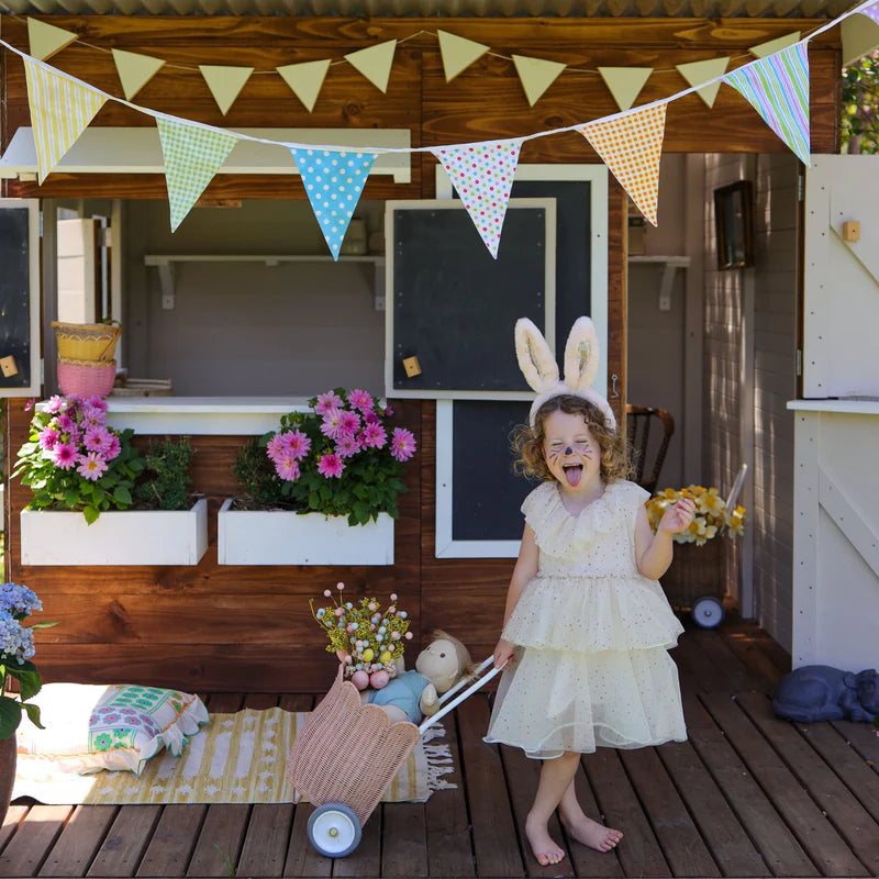 A joyful child in a white dress with the Fluffle Bunny Ear Headband in pink by Olli Ella, surrounded by colorful flowers and bunting, celebrating creativity and imagination.