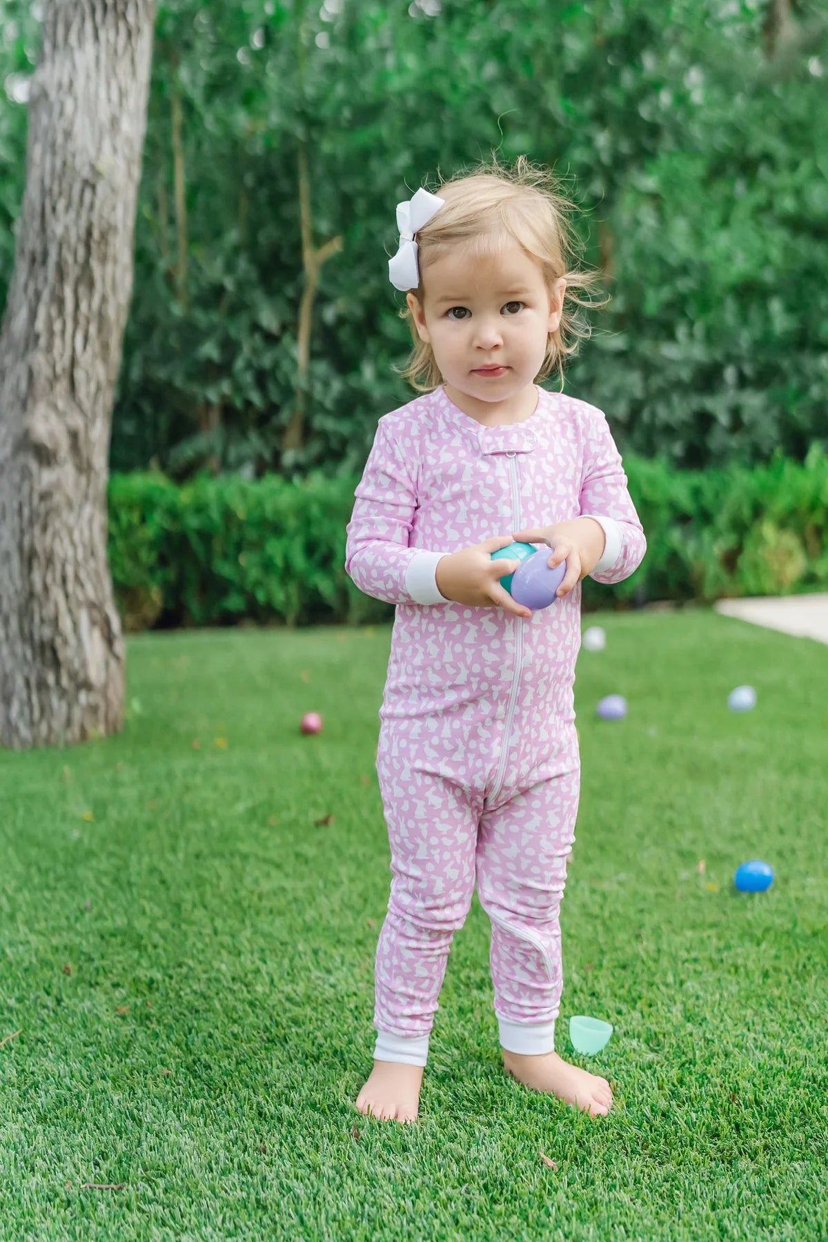 A child wearing the Parker Zipper Pajama Easter Time Pink by Lila and Hayes, holding Easter eggs in a grassy outdoor setting.