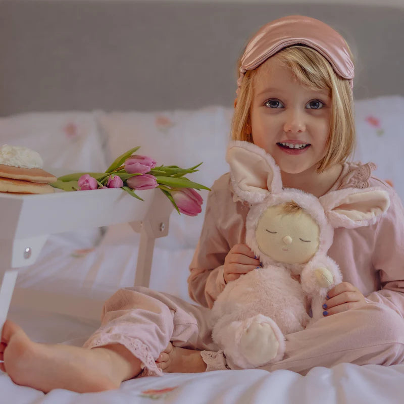 A cheerful girl wearing a pink sleep mask holds the Lullaby Cozy Dinkums Bunny Moppet by Olli Ella in her lap, surrounded by a cozy bedding setup.