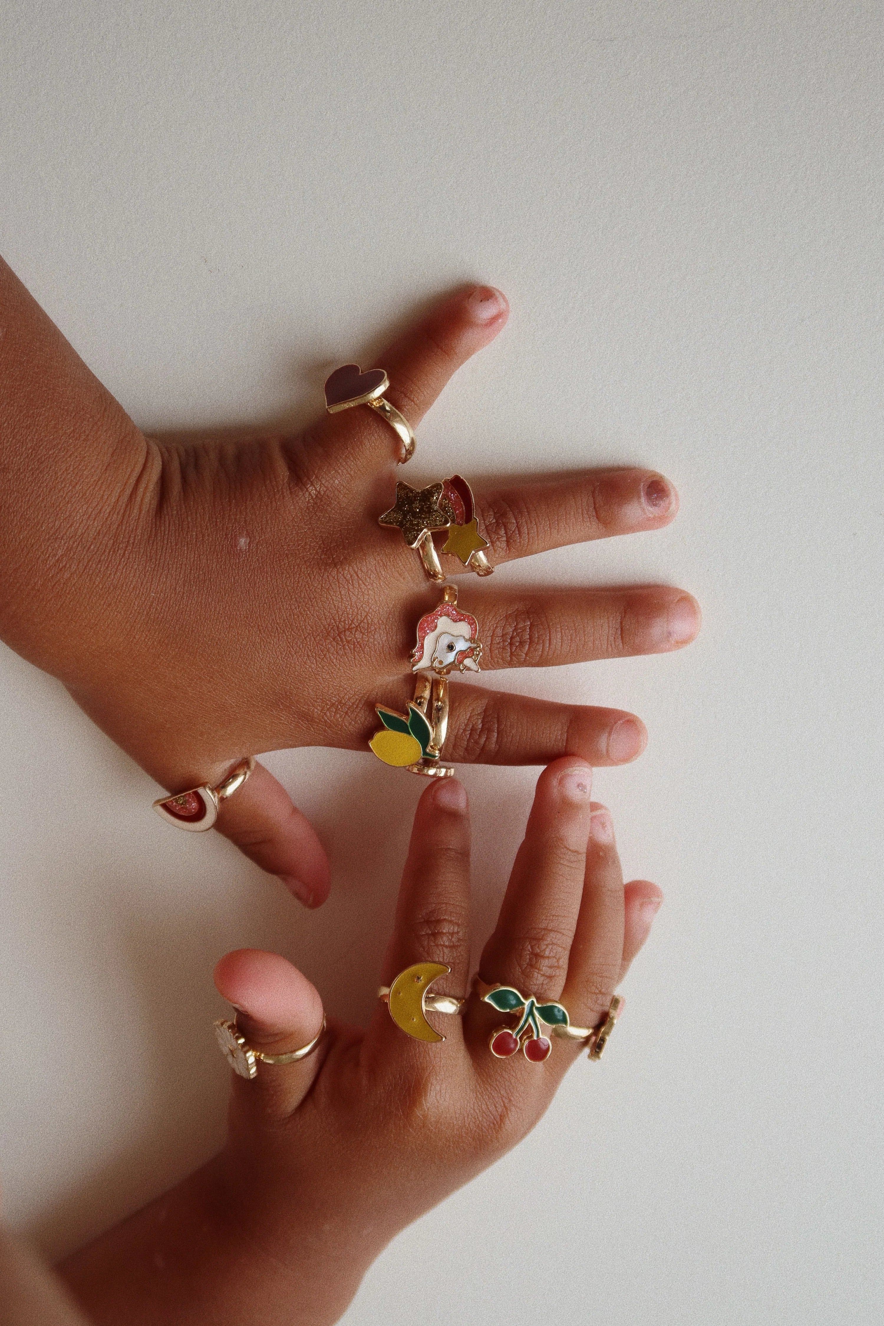 Two hands filled with various rings from the 12 Pack Ring Set from Konges Slojd, each ring showcasing different whimsical motifs.