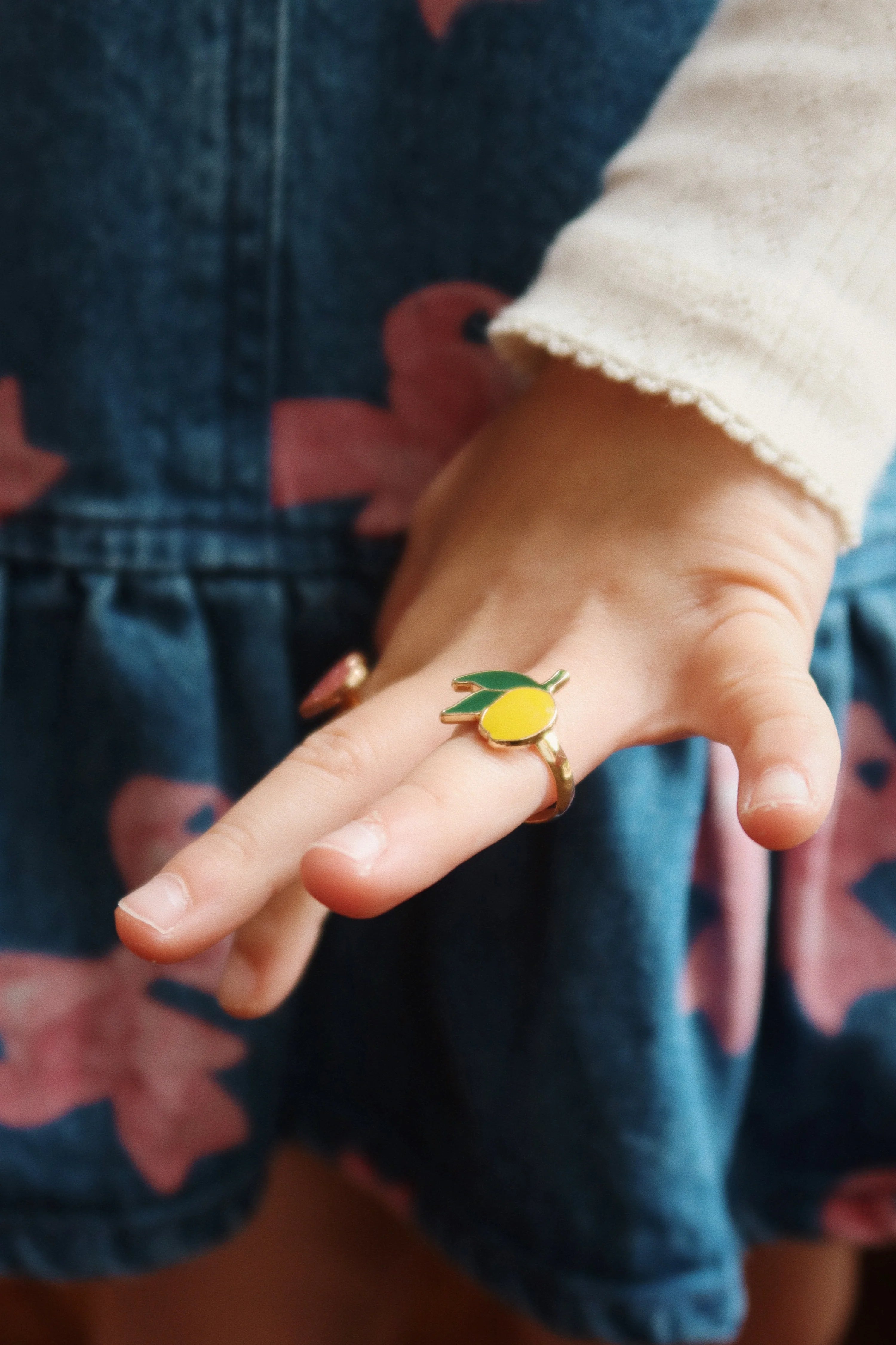 A child’s hand displaying a lemon-themed ring from the 12 Pack Ring Set from Konges Slojd, surrounded by a floral-designed dress.