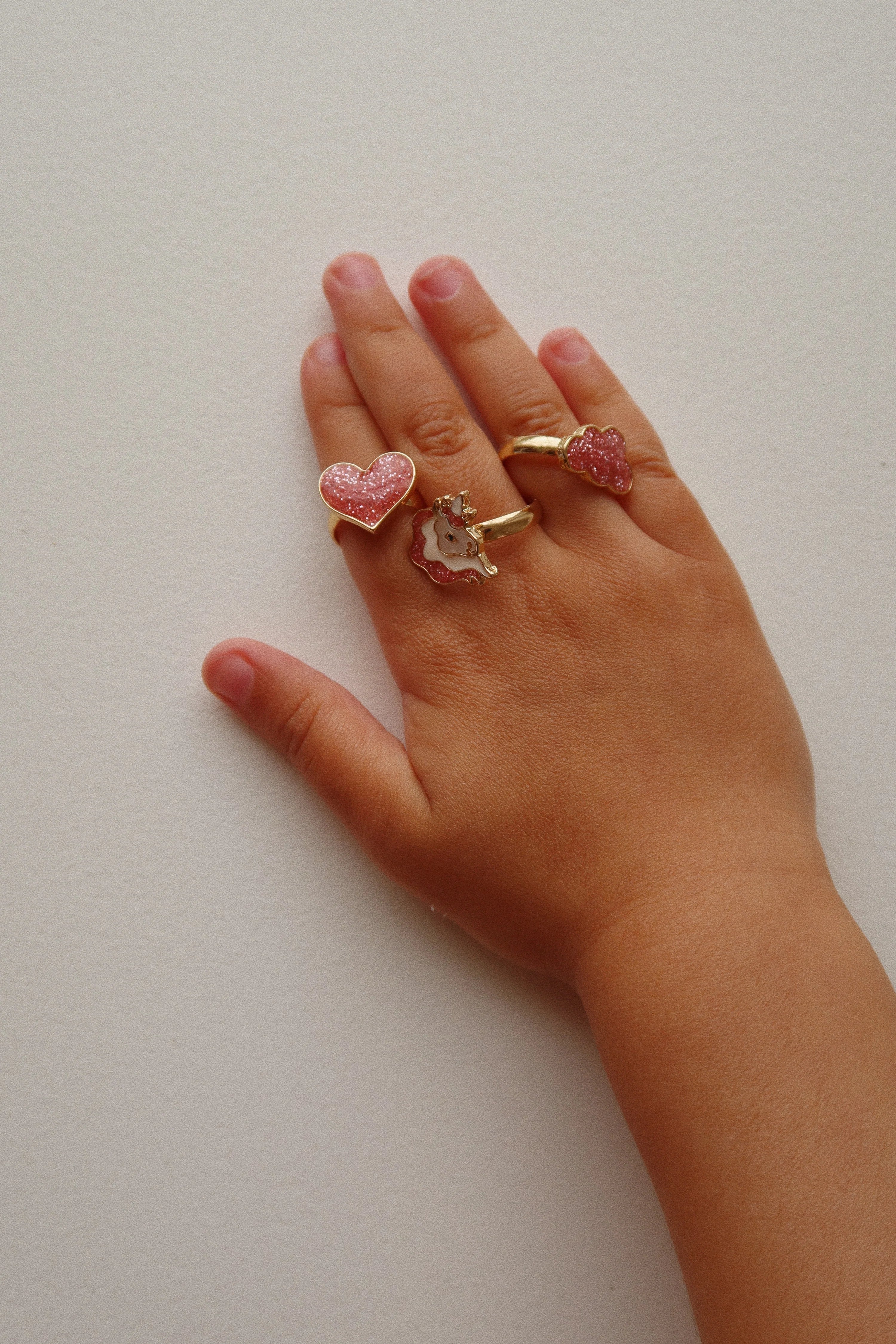 A close-up shot of a child’s hand wearing three different rings from the 12 Pack Ring Set from Konges Slojd, including hearts and unicorn designs.